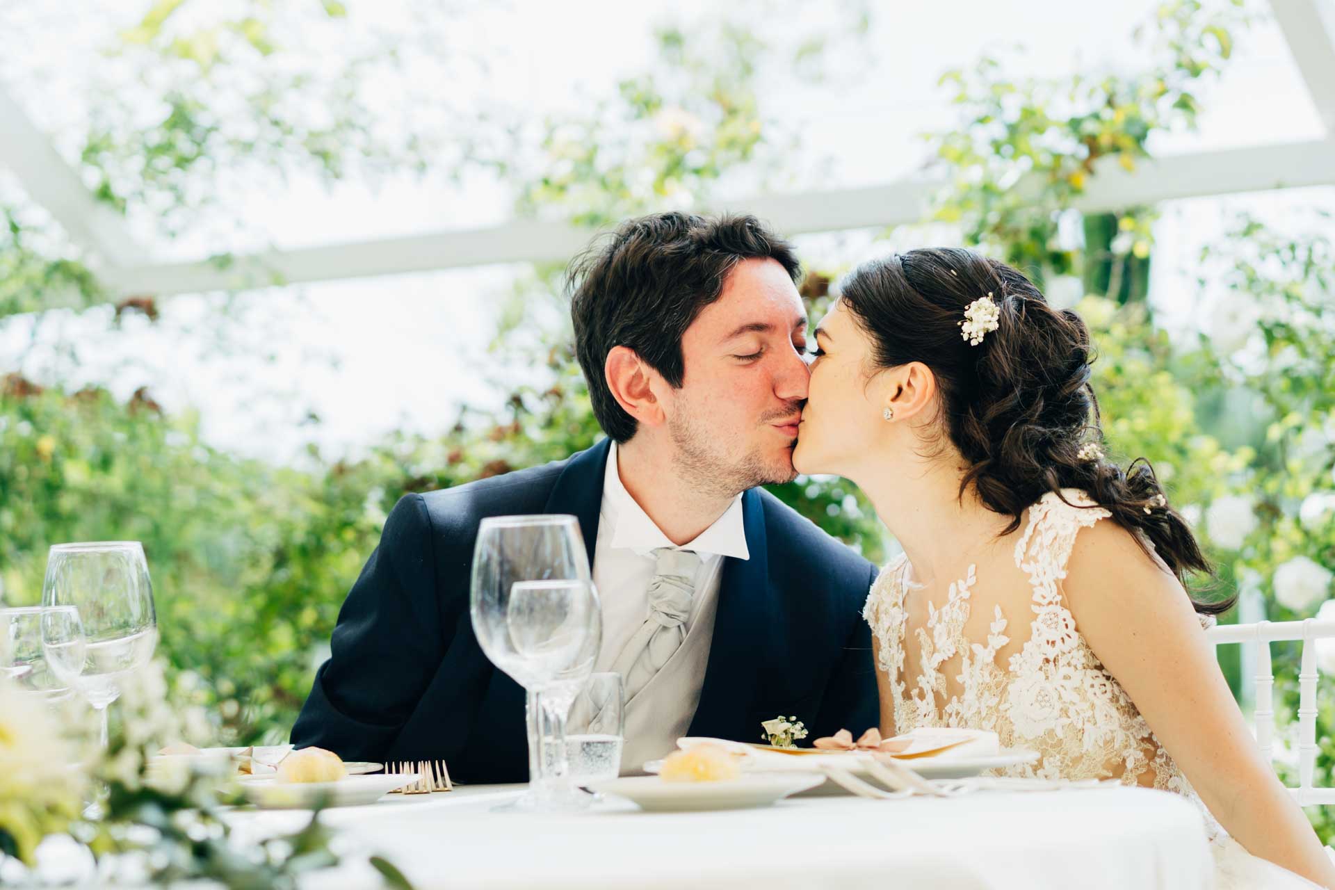 Gli sposi si baciano al tavolo nella sala del ricevimento di matrimonio a Relais Cascina Spiga D'Oro